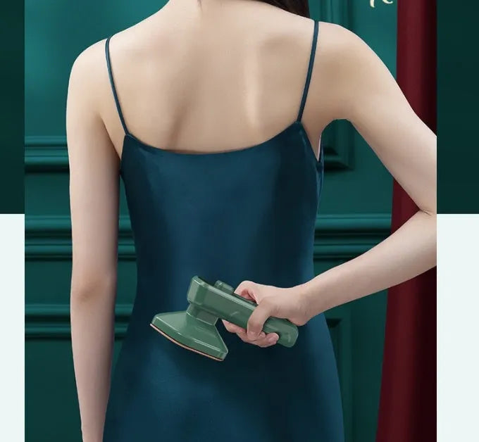 Dark green portable handheld steam iron with ceramic panel, shown next to clothing on a hanger.