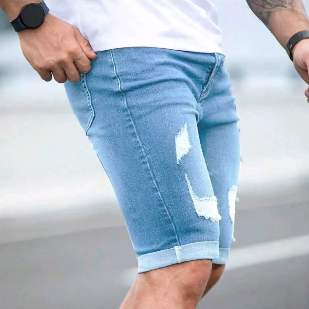 Sky blue men's denim shorts with multi-hole distressing on the legs, showing a low-rise waist and zipper fly.