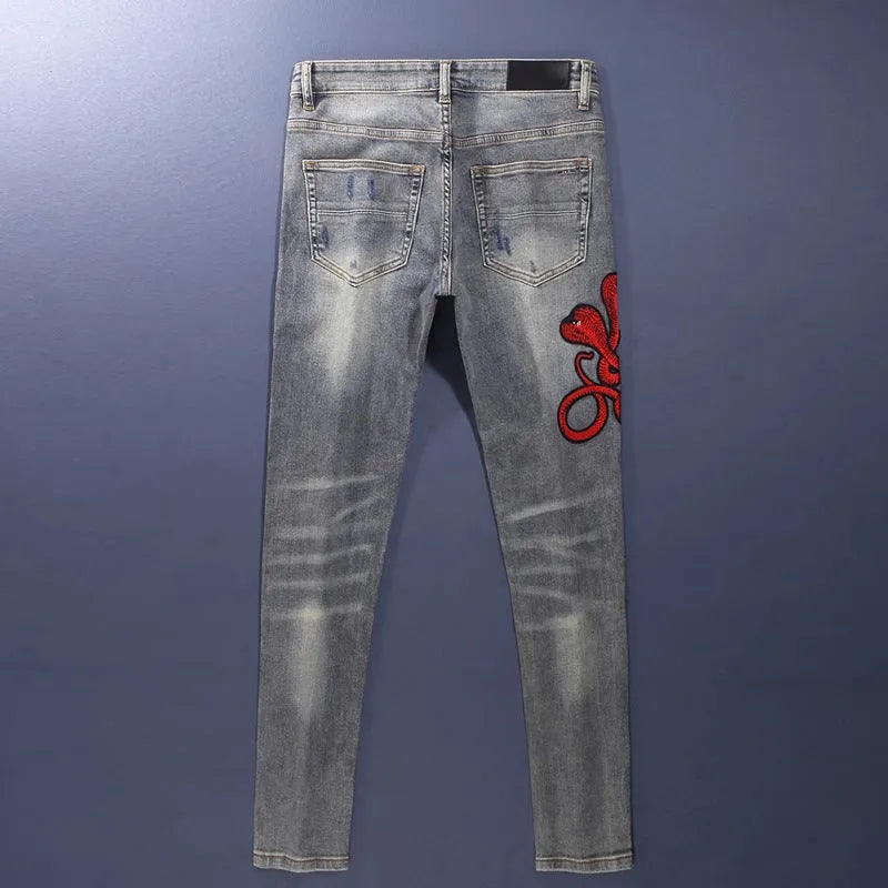 Men's blue slim fit jeans with distressed holes and embroidered patches on faded denim.