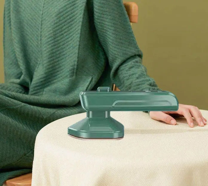 Dark green portable handheld steam iron with ceramic panel, shown next to clothing on a hanger.