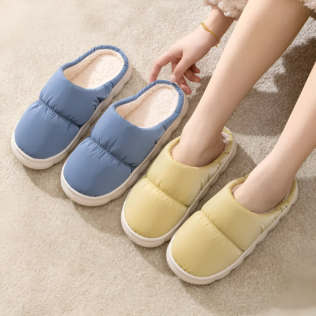 Pair of grey artificial leather slippers with a thick rubber sole and plush lining, shown on a wooden floor.