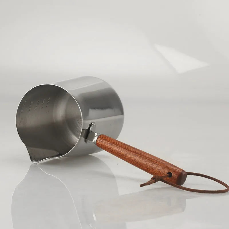 Stainless steel milk pouring cup with wooden handle and interior volume measurement scale.