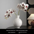White ceramic ball vase with blue stripes and artificial orchid stems on wooden table.