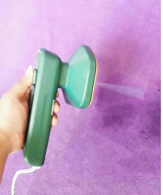 Dark green portable handheld steam iron with ceramic panel, shown next to clothing on a hanger.